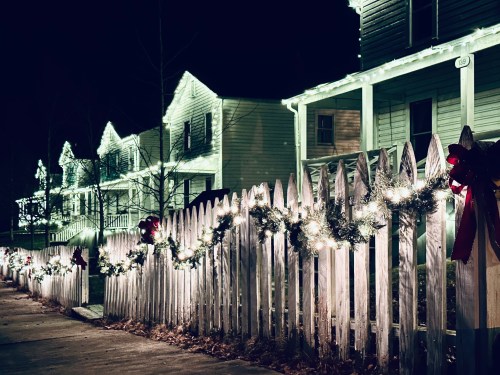A festive night scene of a white picket fence adorned with lights and holiday decorations, with houses in the background.