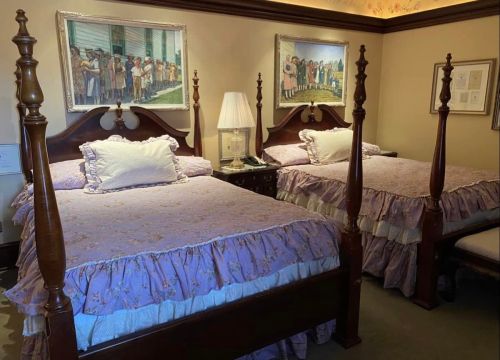 A cozy bedroom with two four-poster beds, each with purple ruffled bedding and framed artwork on the walls.