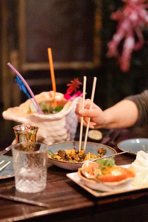 A hand using chopsticks to pick up food from a table with drinks and various dishes in a cozy setting.
