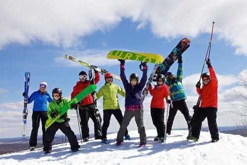 A group of eight skiers and snowboarders pose on a snowy slope, holding their gear and smiling against a blue sky.