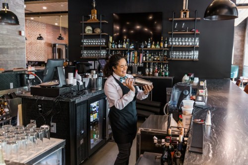 A bartender in an apron shakes a cocktail at a stylish bar with shelves of liquor in the background.