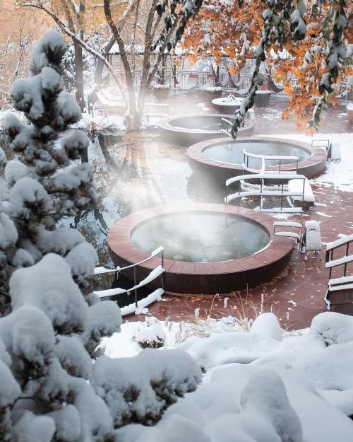 Snow-covered landscape featuring hot tubs surrounded by trees, with steam rising in a serene winter setting.