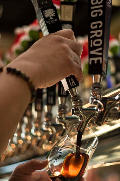 A hand pours a dark beverage from a tap into a glass at a bar with multiple beer taps in the background.