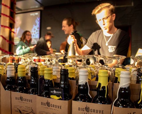 A bartender prepares drinks at a brewery, with bottles in the foreground and patrons in the background.