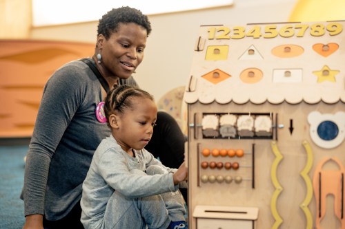 A mother and child engage with a colorful wooden activity board in a playful learning environment.