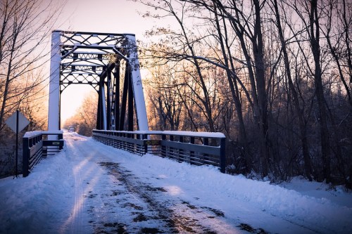 A snow-covered bridge surrounded by trees, with a clear path leading into the distance under a soft winter light.