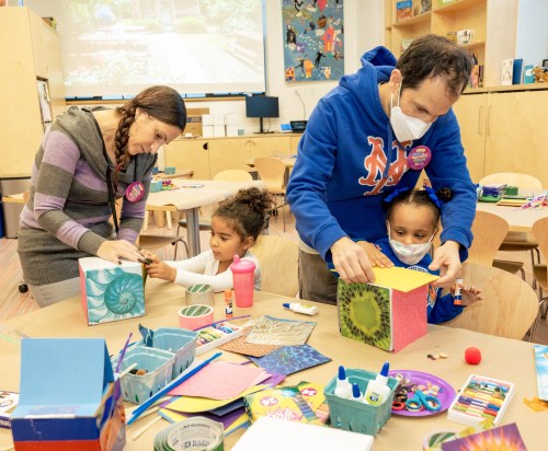 A family engages in a craft activity, decorating colorful boxes with various art supplies in a bright classroom.