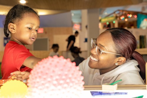 A smiling woman interacts with a young child in a playful, colorful environment filled with toys.
