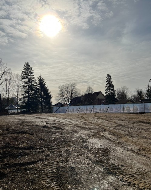 A cleared, muddy lot with construction barriers, trees in the background, and a cloudy sky with the sun peeking through.