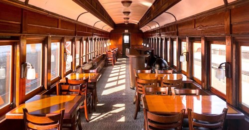Interior of a vintage train car featuring wooden tables, chairs, and large windows with sunlight streaming in.