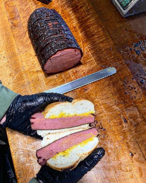 A hand holding a sandwich with sliced meat and mustard, next to a whole smoked meat loaf on a wooden cutting board.