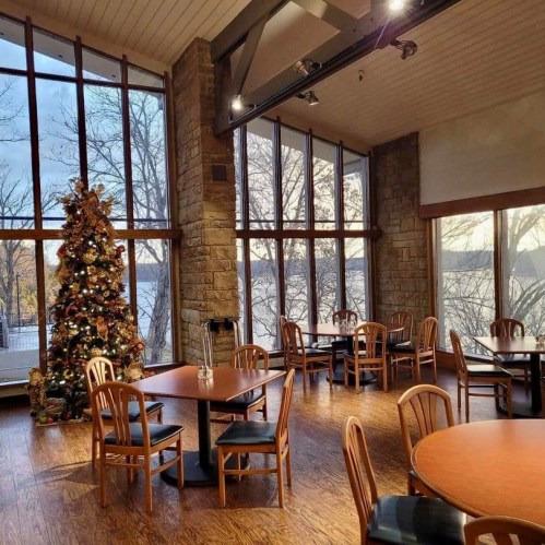 Cozy dining area with wooden tables, chairs, a Christmas tree, and large windows overlooking a scenic view.