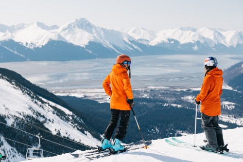 Two skiers in orange jackets stand on a snowy mountain, overlooking a valley and distant snow-capped peaks.