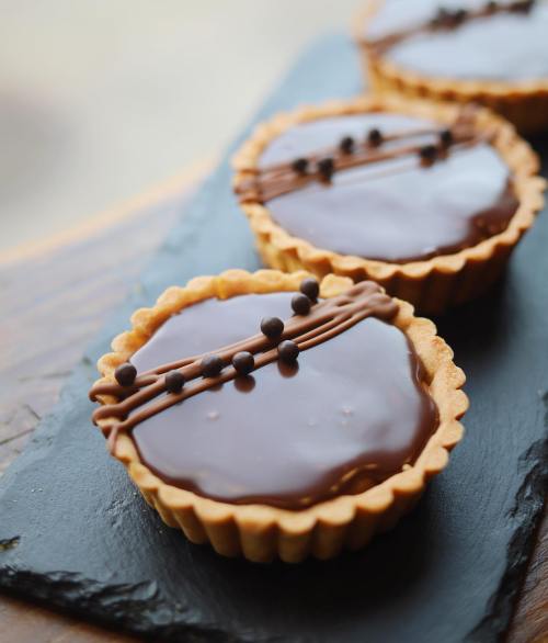 Three chocolate tarts with a glossy topping and decorative chocolate drizzle, placed on a dark slate surface.