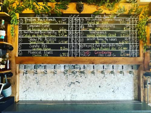 A chalkboard menu displaying various drinks and prices at a bar, surrounded by greenery.