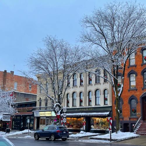 A snowy street scene featuring historic buildings, bare trees, and a parked car.