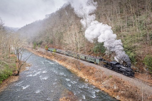A steam train chugs along a riverbank, surrounded by misty mountains and bare trees. Smoke billows from the engine.