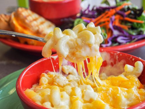 A close-up of creamy macaroni and cheese with melted cheese, served in a red bowl, with salad and grilled bread in the background.