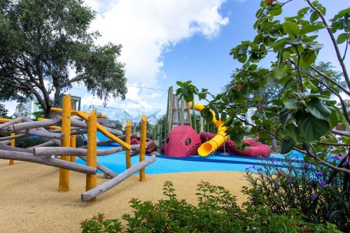 Colorful playground with slides and climbing structures, surrounded by greenery and a blue rubber surface.