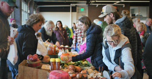 A bustling farmers market with people shopping for fresh produce and goods at various vendor tables.