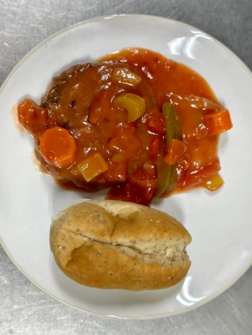 A plate with a meat patty in tomato sauce with vegetables, accompanied by a small brown bread roll.