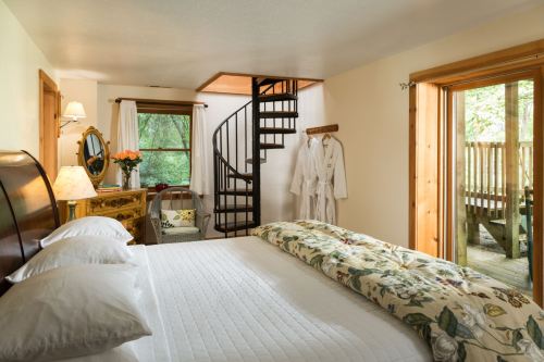 Cozy bedroom with a spiral staircase, wooden furniture, and a door leading to a deck surrounded by greenery.