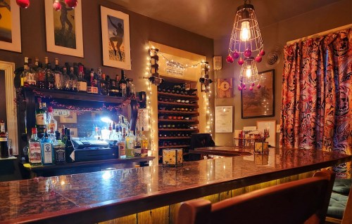 Cozy bar scene with a marble countertop, shelves of liquor, wine racks, and warm lighting. Colorful curtains add charm.