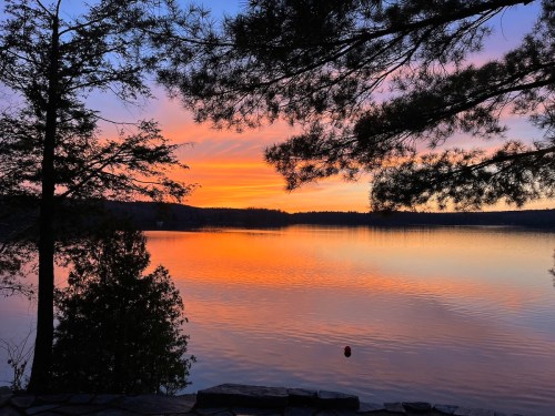 A serene lake at sunset, reflecting vibrant orange and purple hues, framed by trees on the shore.