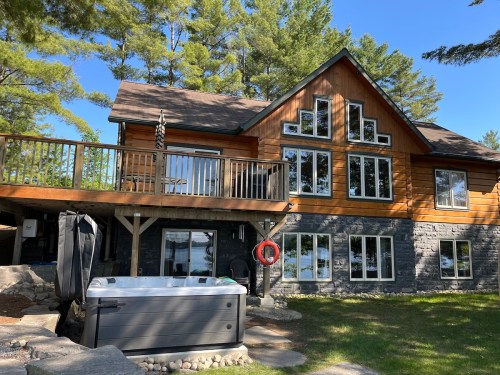 A lakeside cabin with large windows, a wooden deck, and a hot tub in the foreground, surrounded by trees.
