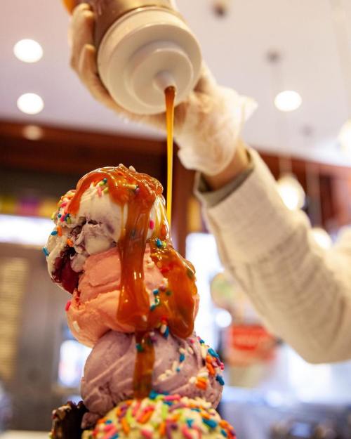 A hand drizzles caramel sauce over a tall stack of colorful ice cream scoops topped with sprinkles.