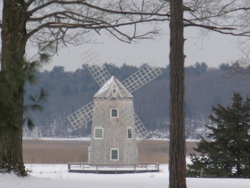 A snow-covered landscape featuring a wooden windmill surrounded by trees and a distant forest.