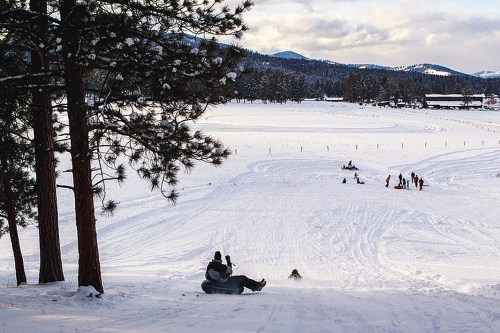 A snowy landscape with people sledding down a hill, surrounded by trees and mountains in the background.