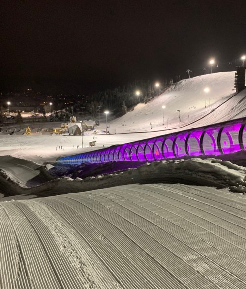 A snowy ski slope at night, featuring a brightly lit, purple-tinted tunnel and skiers in the distance.