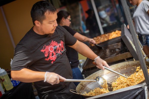 A chef in a black shirt stirs fried rice in a large pan at a food stall, with others preparing food in the background.
