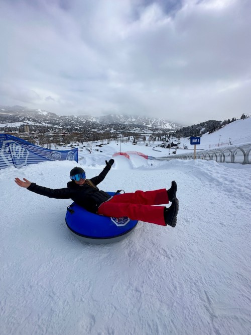 A person in a blue helmet and red pants joyfully tubing down a snowy slope, with mountains in the background.