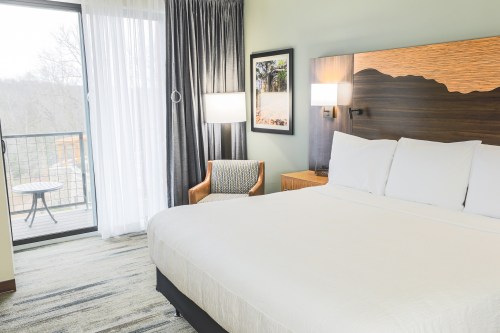 A cozy hotel room featuring a king-sized bed, a chair, and a balcony with a view. Natural light fills the space.