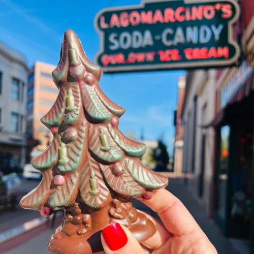 A hand holds a chocolate Christmas tree in front of a vintage soda candy shop sign.