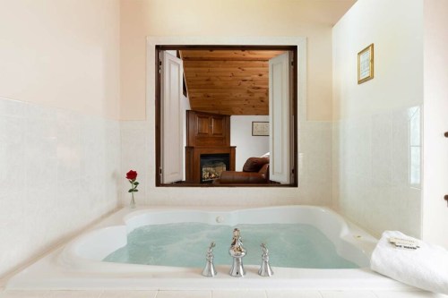 A luxurious bathtub with water, a rose, and a view of a cozy room with a fireplace through an open window.