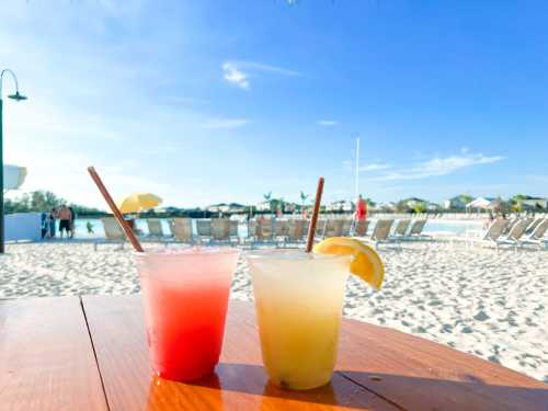 Two colorful cocktails on a wooden table, with a beach and lounge chairs in the background under a clear blue sky.