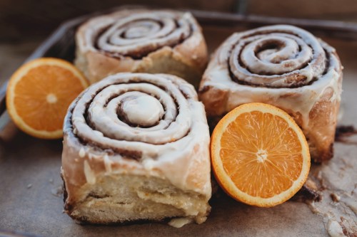 Three cinnamon rolls with icing, placed on a baking tray alongside two orange halves.