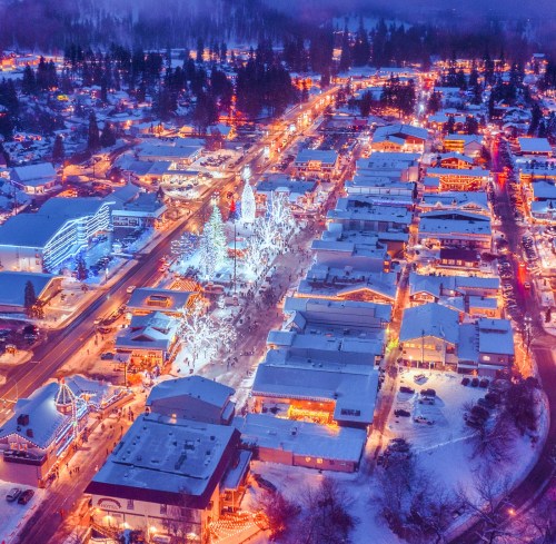 Aerial view of a snowy town illuminated with colorful holiday lights, featuring decorated trees and bustling streets.