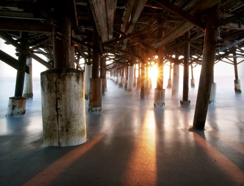 Sunlight streams through the wooden supports of a pier, casting reflections on the water below.