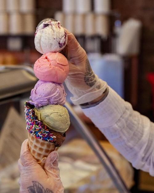 A hand in a glove holds a tall ice cream cone with four colorful scoops and sprinkles on top.