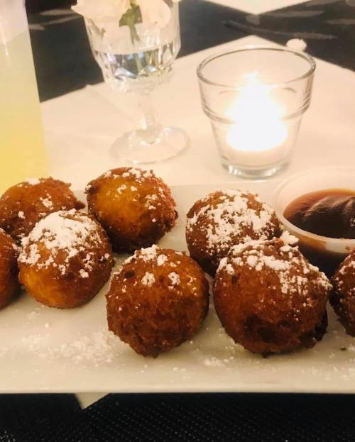 A plate of golden-brown fried dough balls dusted with powdered sugar, served with a small bowl of sauce.