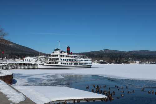 A large steamboat docked by a frozen lake, surrounded by snow-covered mountains under a clear blue sky.