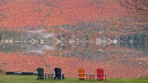 Four colorful Adirondack chairs sit by a lake, reflecting vibrant autumn foliage and a serene landscape.