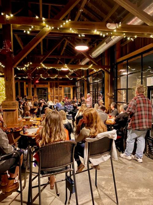 A lively gathering in a rustic venue, with people seated and a speaker at the front, illuminated by string lights.