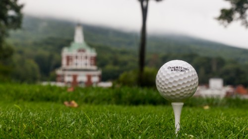 A golf tee with a ball in focus, set against a blurred background of a historic building and misty mountains.