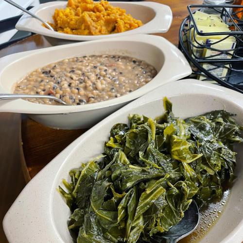 Three dishes on a table: black-eyed peas, sweet potato mash, and collard greens in serving bowls.
