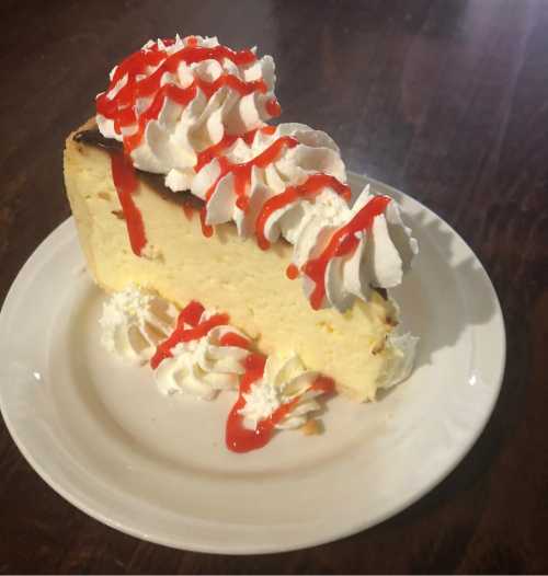 A slice of cheesecake topped with whipped cream and drizzled with red syrup, served on a white plate.